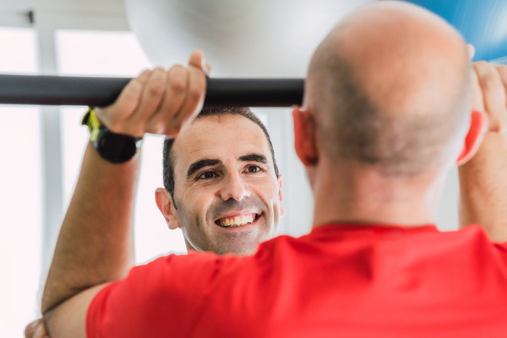A smiling fitness trainer coaches a man through a barbell shoulder press.