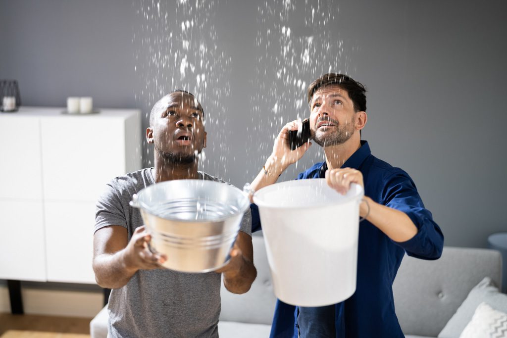 Two men in a gym hold buckets to catch water raining from a leaky roof.
