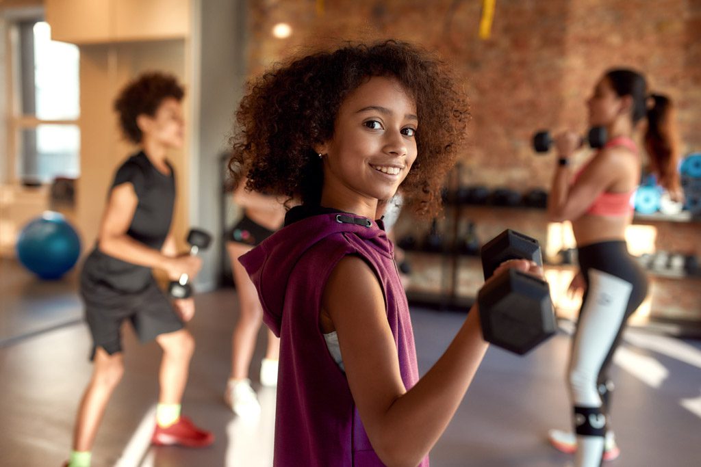 A teenager performs a biceps curl with a dumbbell in a gym, with her coach and other kids in the background.