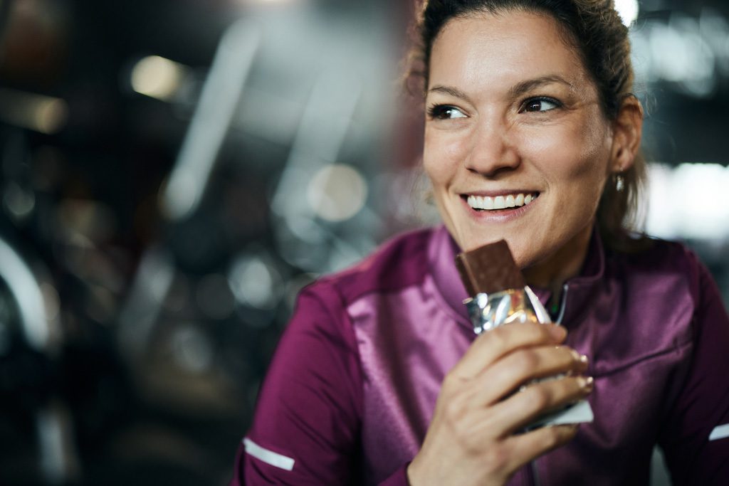A smiling woman in a purple top eat a protein bar after a training session in a gym.