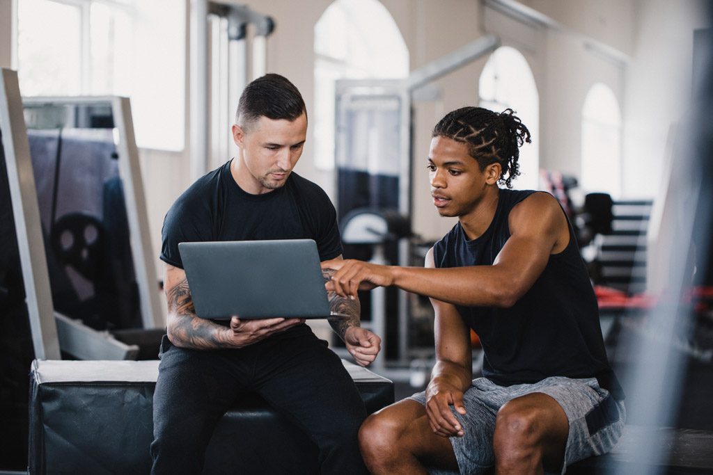 A young athlete in a gym reviews his progress toward goals with a trainer who is holding a laptop.