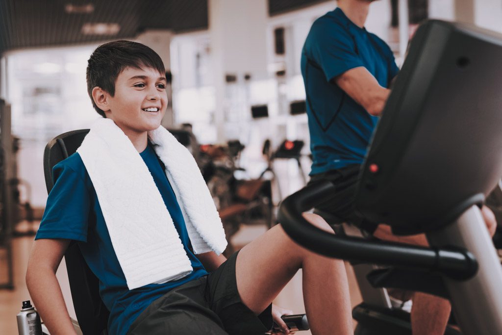 A teenaged boy smiles as he rides an exercise bike in a large fitness facility.