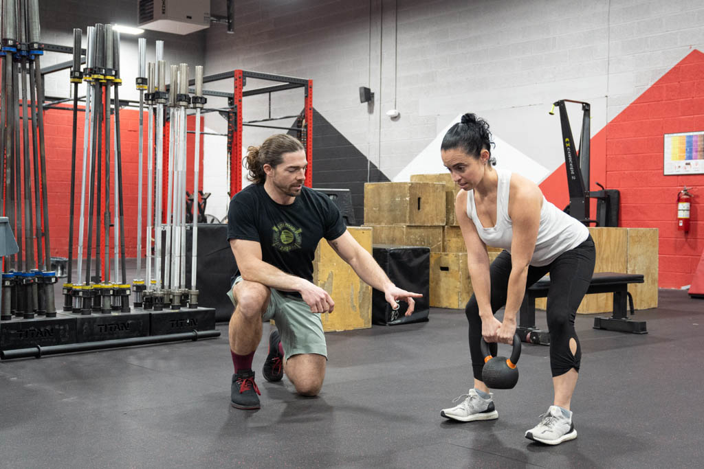 In a functional fitness gym, a trainer coaches a woman through kettlebell deadlifts designed to increase strength.