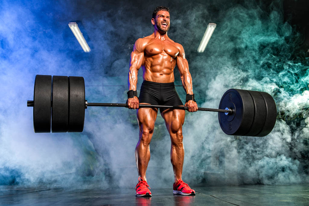 A muscular, shirtless man in black shorts performs a heavy barbell deadlift.