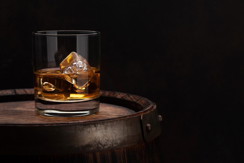 A close-up image of a glass of whiskey and ice on top of an oak barrel.
