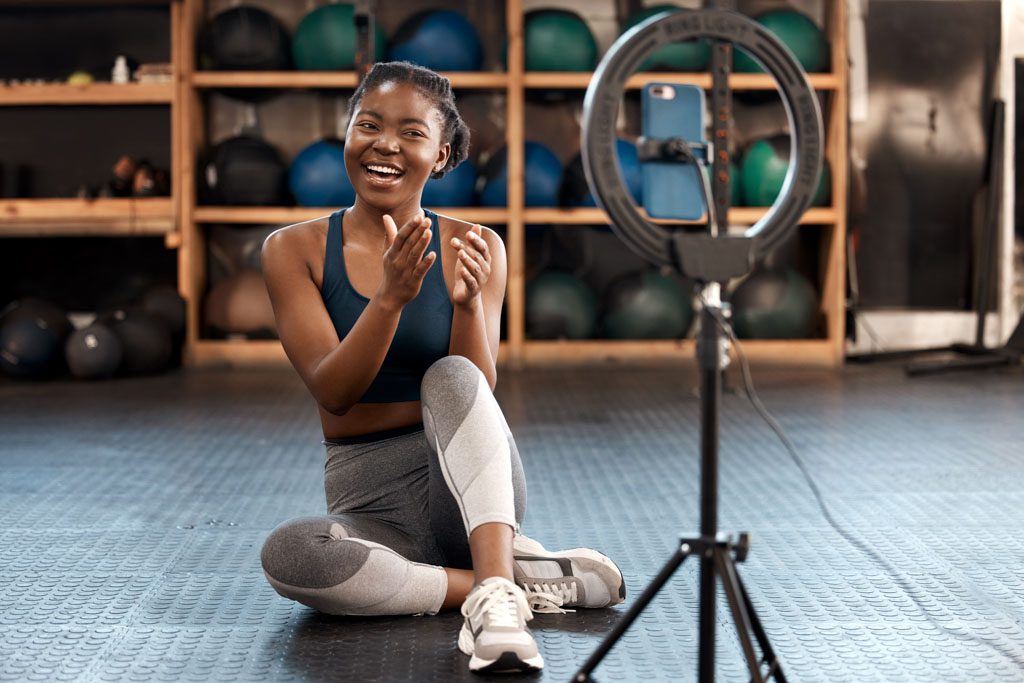 A gym clients sits and uses a ring-light tripod and smartphone to create a video about her workout.