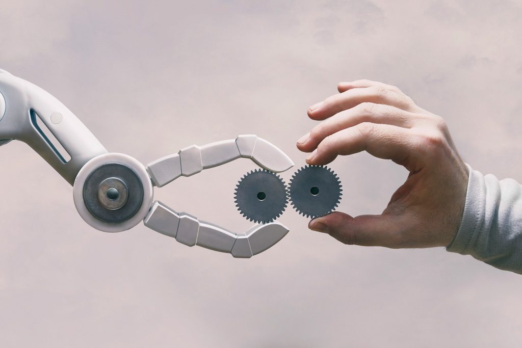 A human hand holding a small gear connects the gear to another held by a robotic arm.
