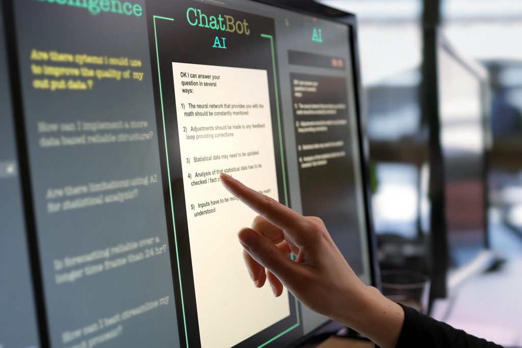 Close-up photo showing a touchscreen monitor: A woman’s hand is typing questions and an AI chatbot is answering.