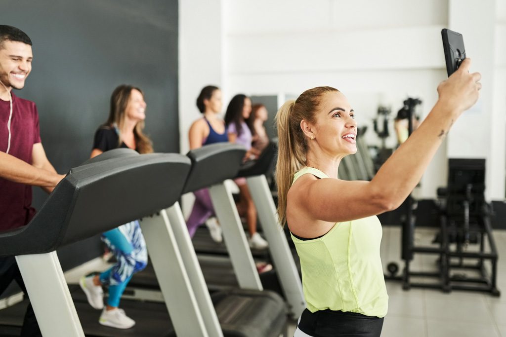 A smiling gym owner takes a selfie with happy clients on treadmills behind her.