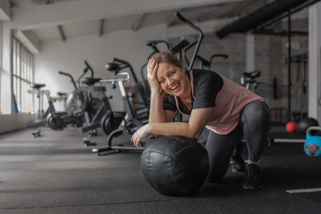 A woman with a surprised look on her face rests on a medicine ball in a functional fitness gym.