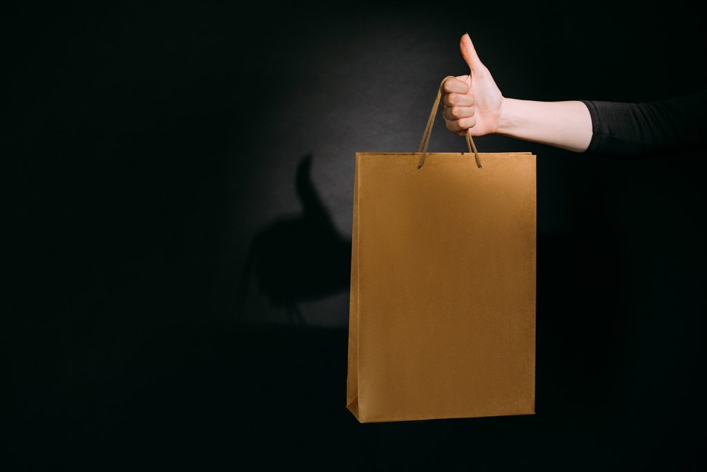 Unrecognizable woman holding beige present bag in hand and making thumbs-up gesture.
