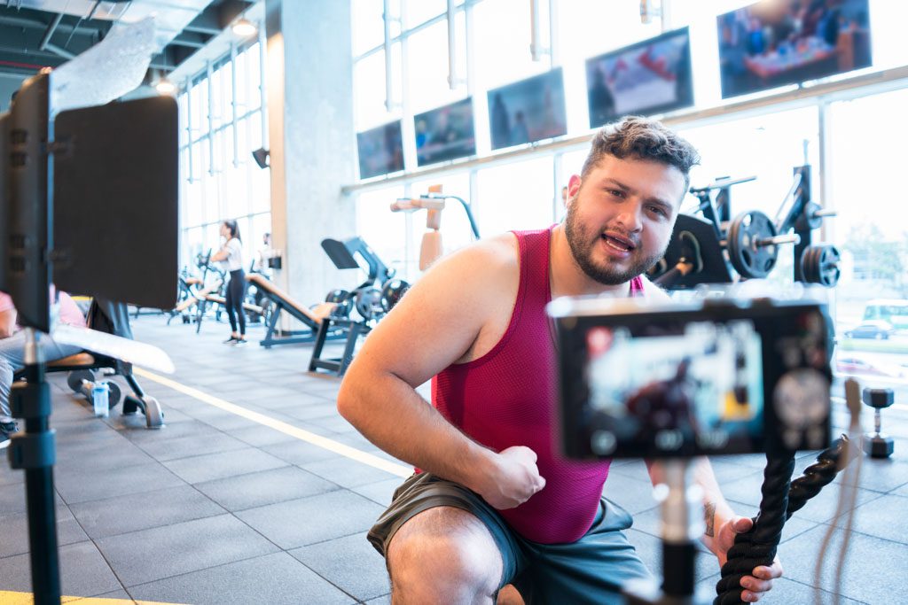 Average age man dressed in a sporty way is in front of his camera in a gym to record a training video for his fans on YouTube.