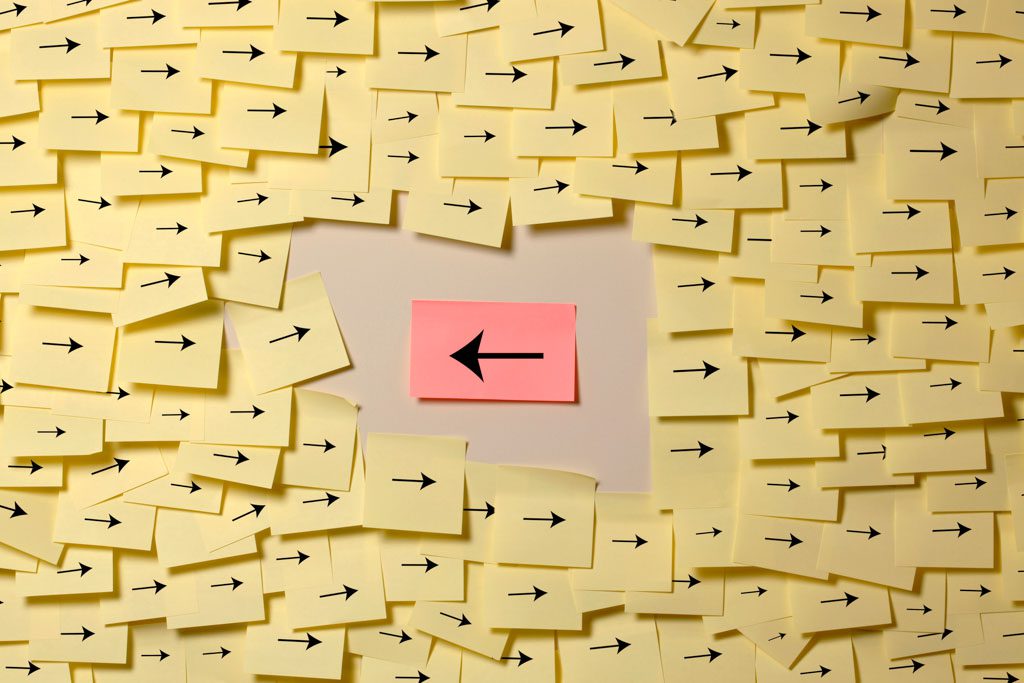 In a sea of yellow sticky notes with arrows pointing to the right, one pink note in the center has an arrow pointing left.