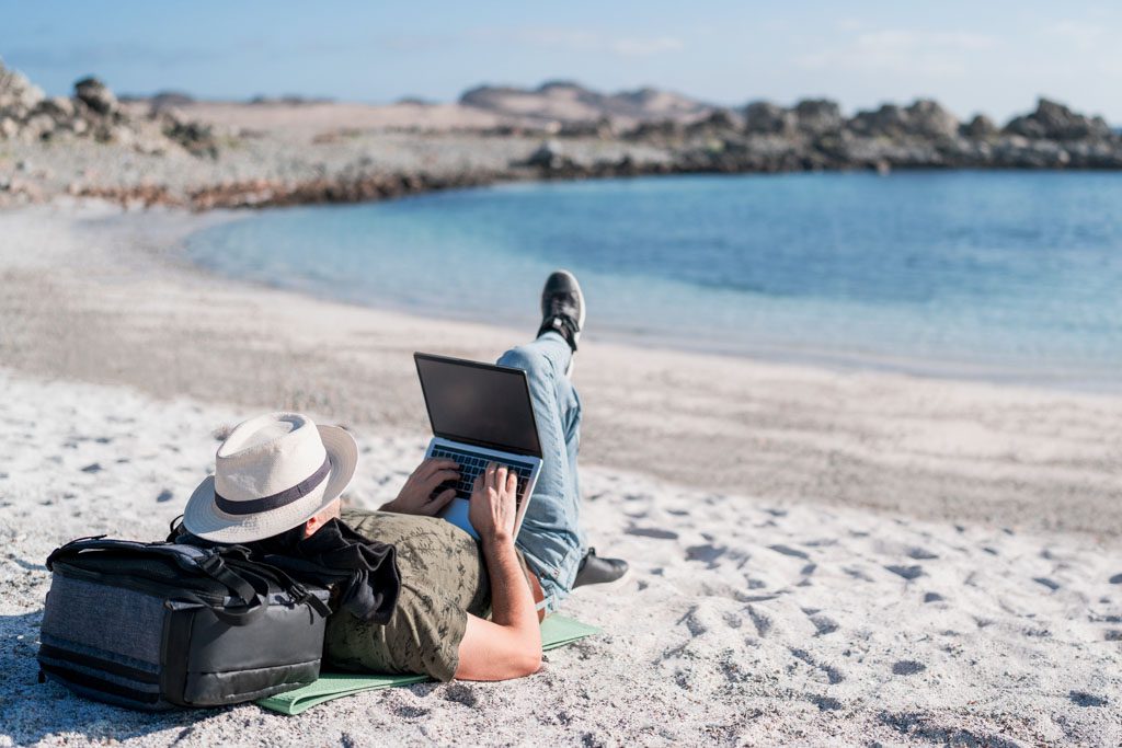 A fitness entrepreneur reclines on a beach with his head on a backpack as he uses a laptop to check in.