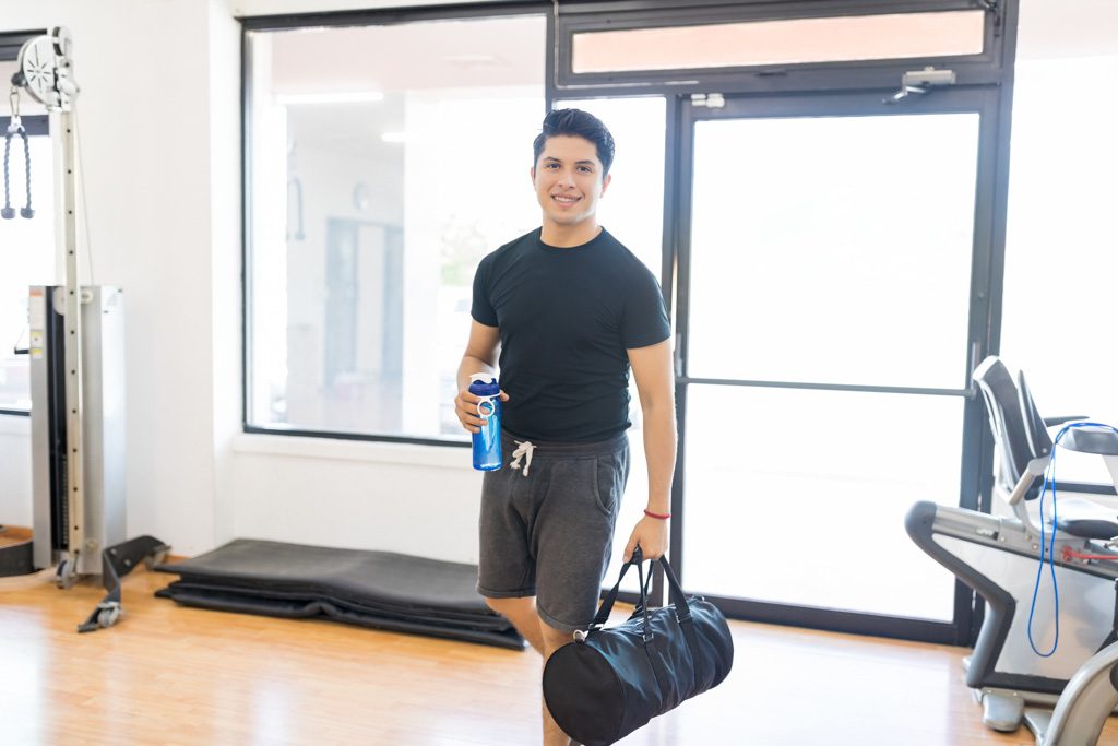 Smiling, fit man with water bottle and gym bag entering health club.