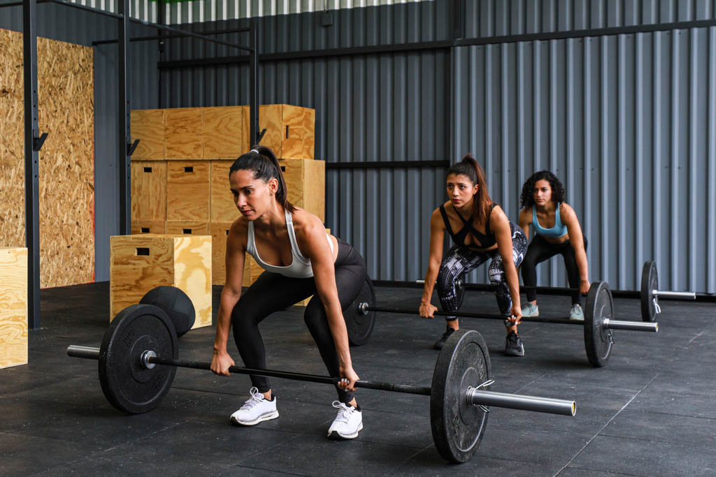 Three women grip barbells at the bottom of the deadlift position before starting a fitness competition.