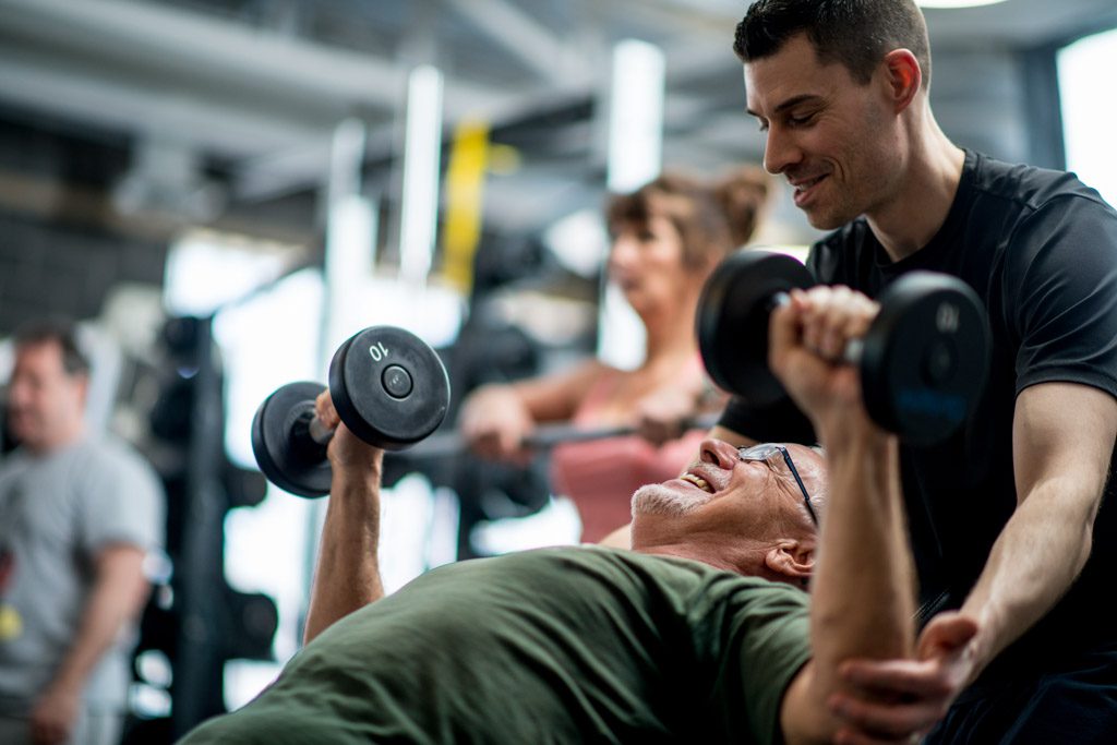 A smiling personal trainer helps and older client perform dumbbell chest presses.