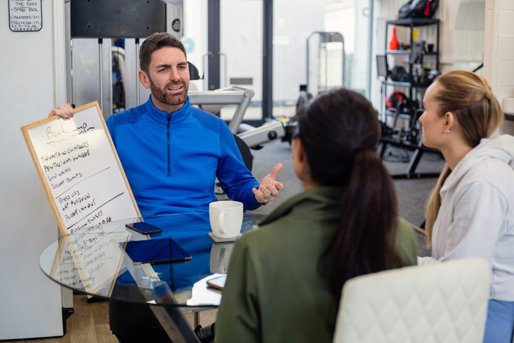 A gym owner sits at a table and does a free fitness consultation with two prospective clients.