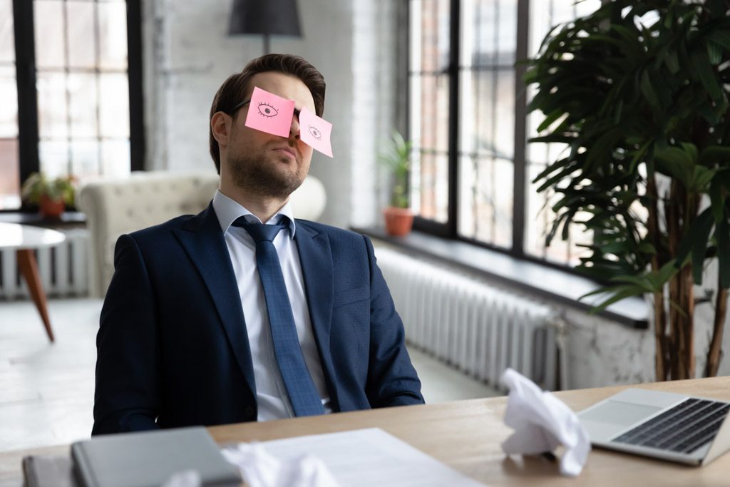A CEO sleeps at his desk with open eyes drawn on sticky notes stuck to his forehead.