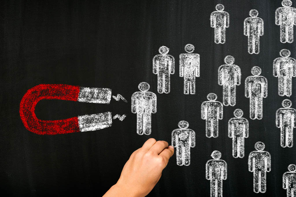 On a chalk board, a hand draws people icons being attracted to a large, red magnet.