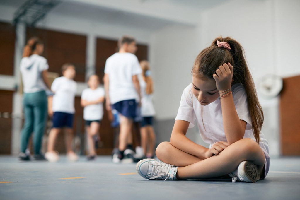A young athlete sits on the floor and sulks while peers perform fitness activities behind her.