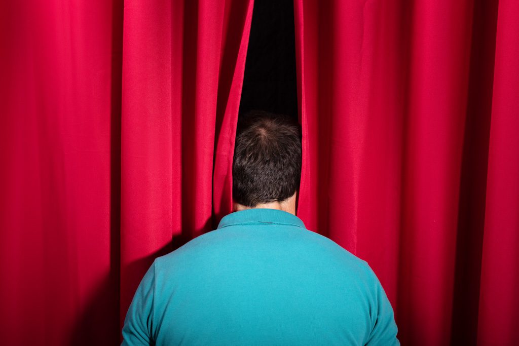 A graphic showing a person peeking behind a bright red stage curtain.