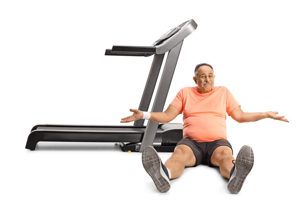 A confused man sits on the floor next to a treadmill at a gym.