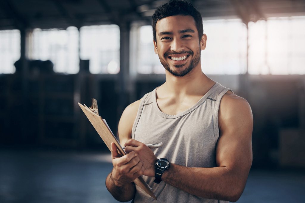 A smiling personal trainer holds a clipboard in a gym.