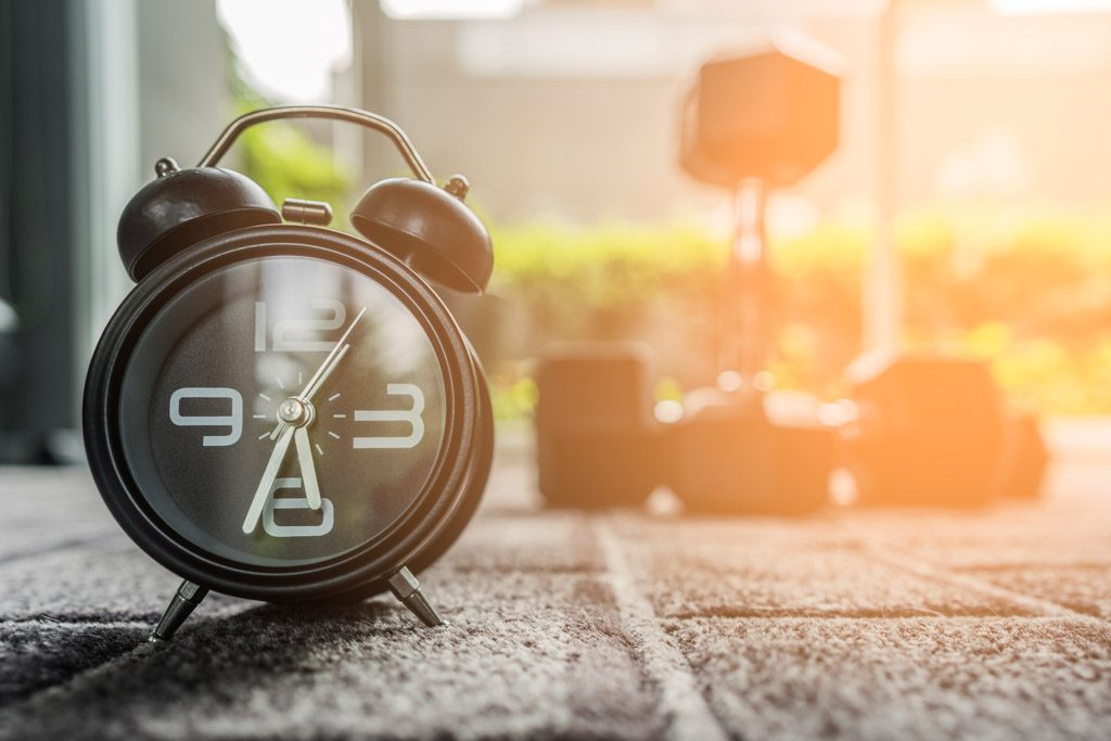 An alarm clock sits on the floor of a gym, with a stack of dumbbells in the background.