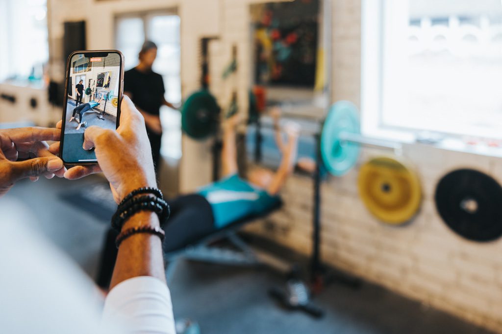 A gym owner uses a cell phone to film a trainer coaching a client to perform bench presses.