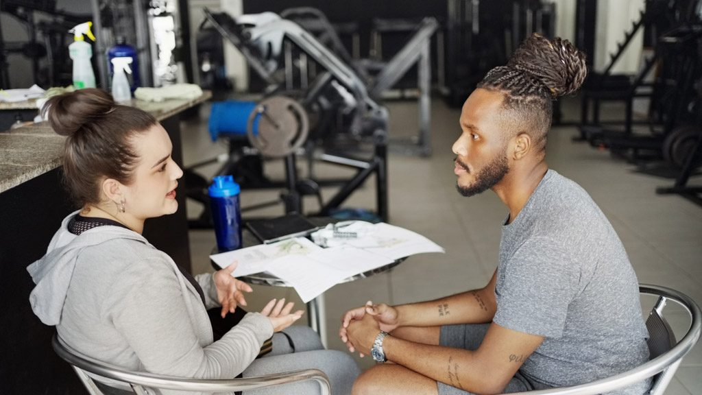 A man sits in a gym and consults with its owner about his fitness goals.