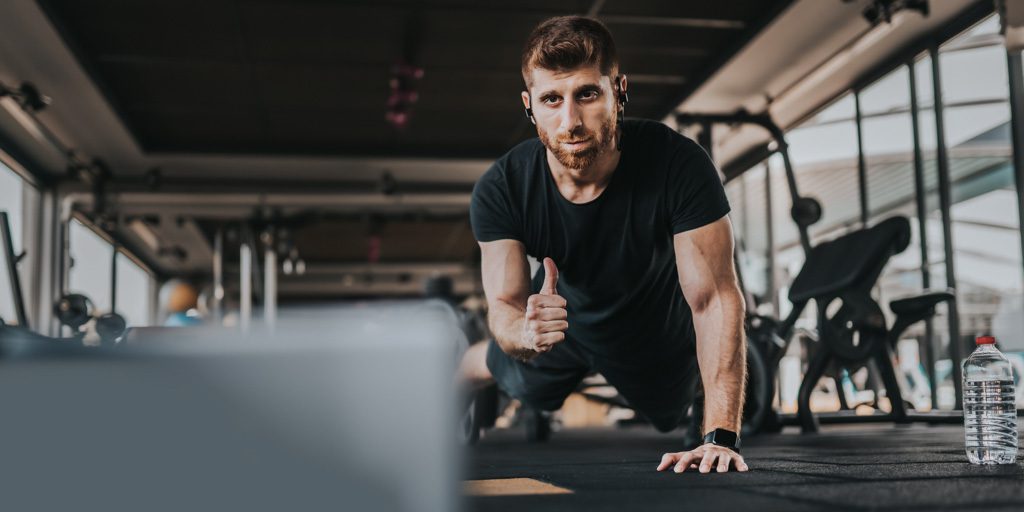 Personal trainer/fitness coach in a gym gives a thumbs up mid-push up while looking at his computer