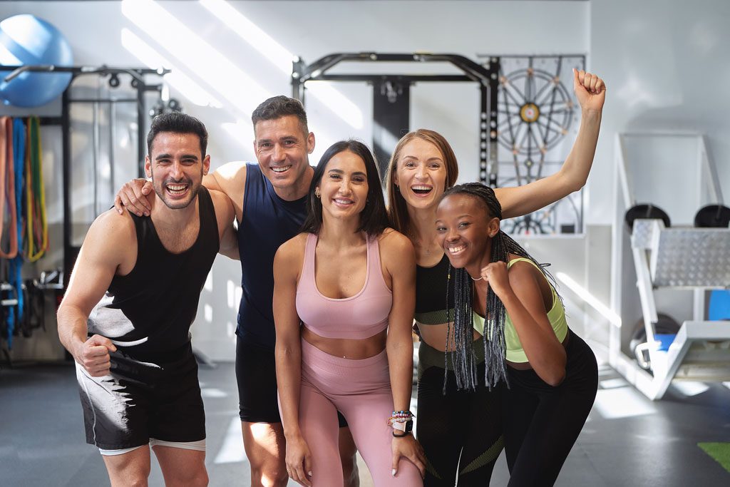 A small group of 5 fitness clients smile as they pose in front of equipment in a gym.