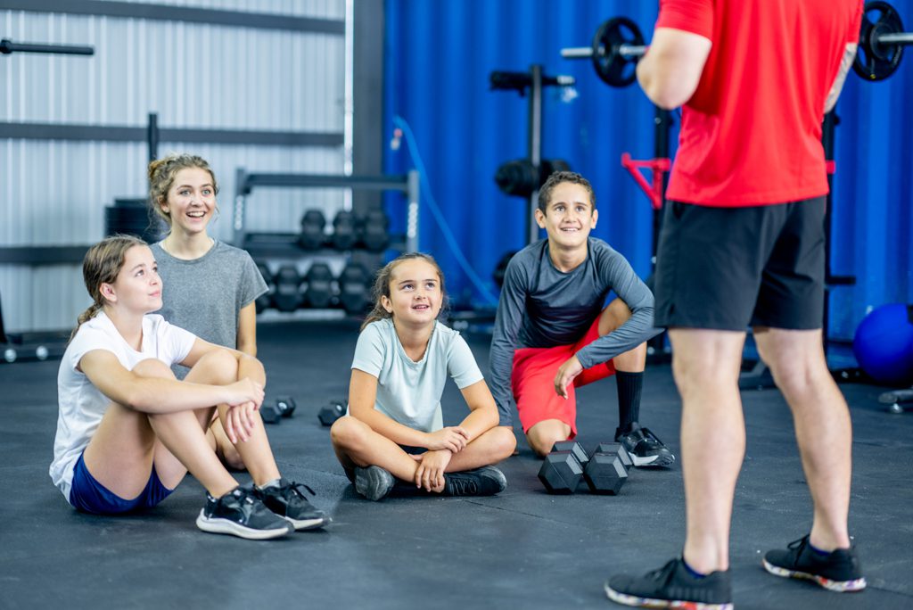A group of four children listen to a coach as part of a summer fitness program in a gym.