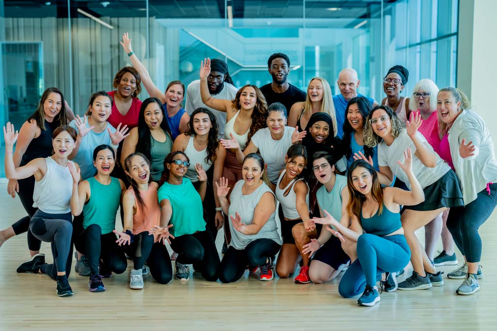 A large group of gym clients smile and wave after a fitness class.