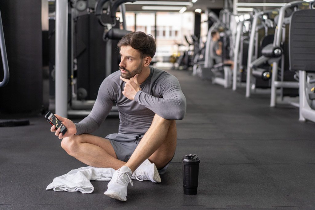 A gym owner sits on the floor reading from his phone instead of working out.