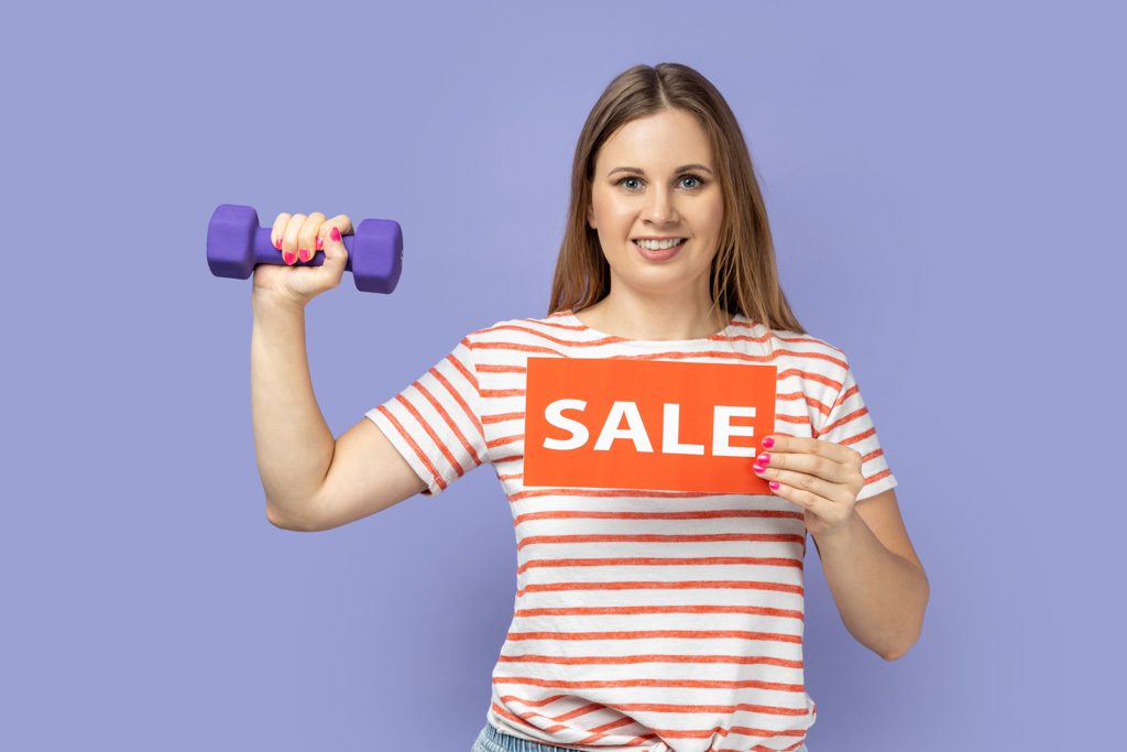 A woman holds up a small purple dumbbell and an orange card that reads "sale."