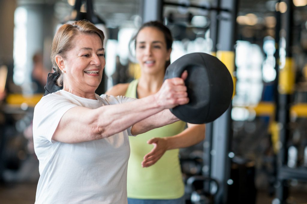 A personal trainer coaches a woman through a front delt raise with a medicine ball.