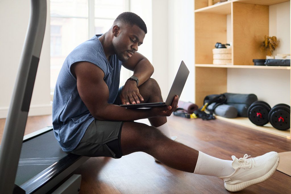 A gym owner sits on a treadmill and reviews financial statements on a laptop.