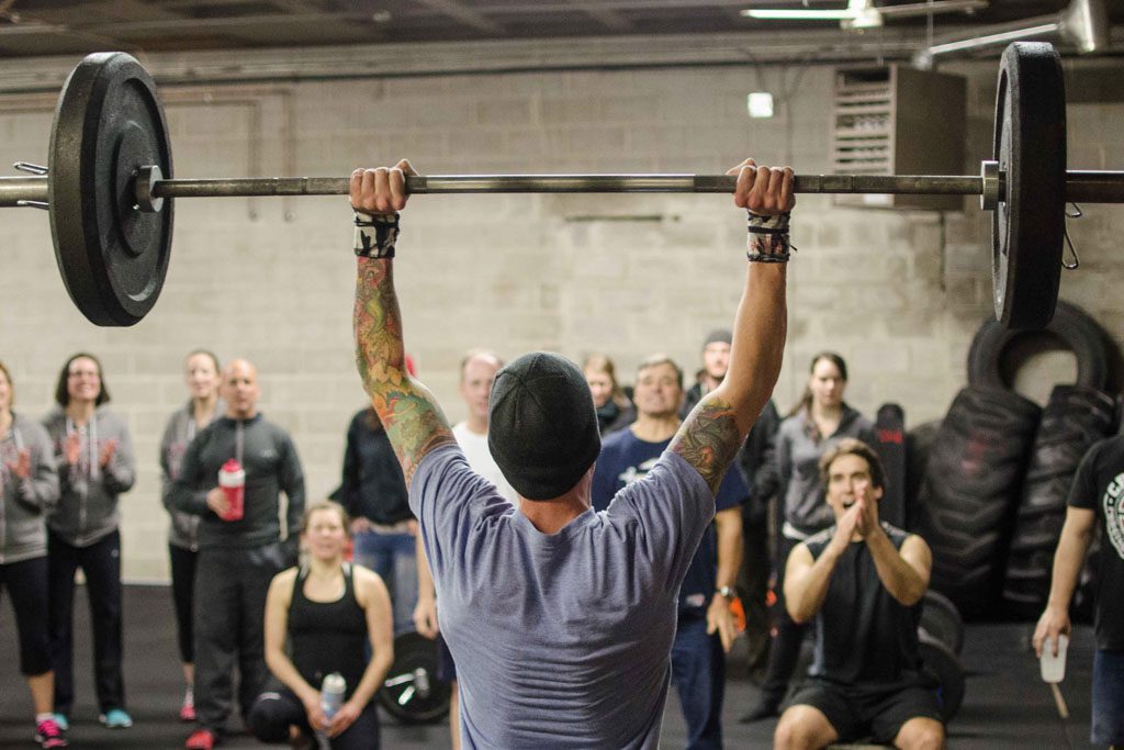 A crowd cheers an athlete on as he does barbell thrusters in a gym.