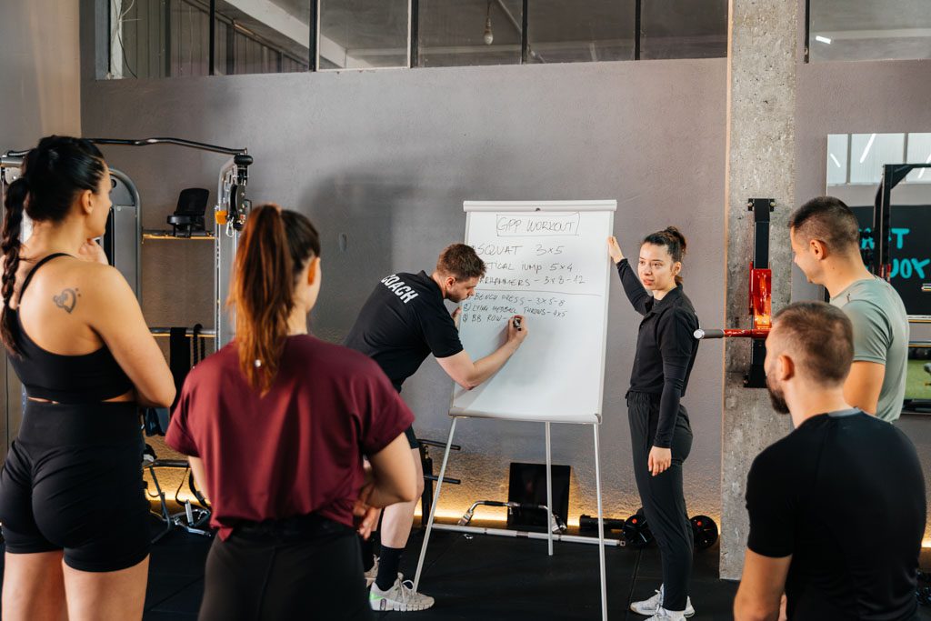 On a whiteboard, a coach presents a workout to a small group of clients at a gym.