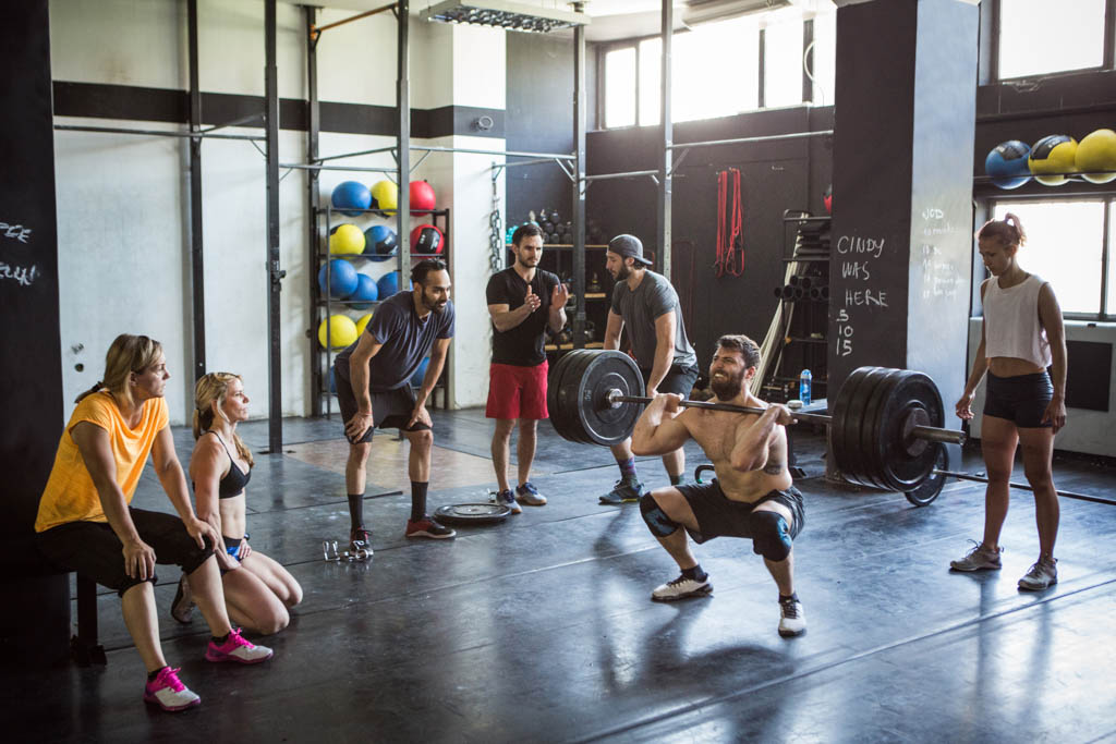 A group of people watch a man perform a barbell clean in a functional fitness gym.