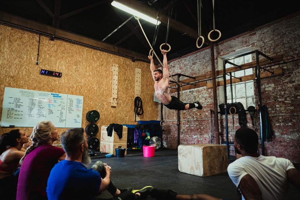 A man does a ring muscle-up in a gym while other people evaluate his technique.