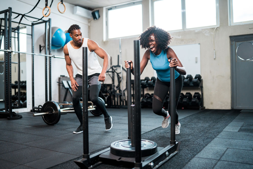 A personal trainer coaches a client through sled pushes in a gym.
