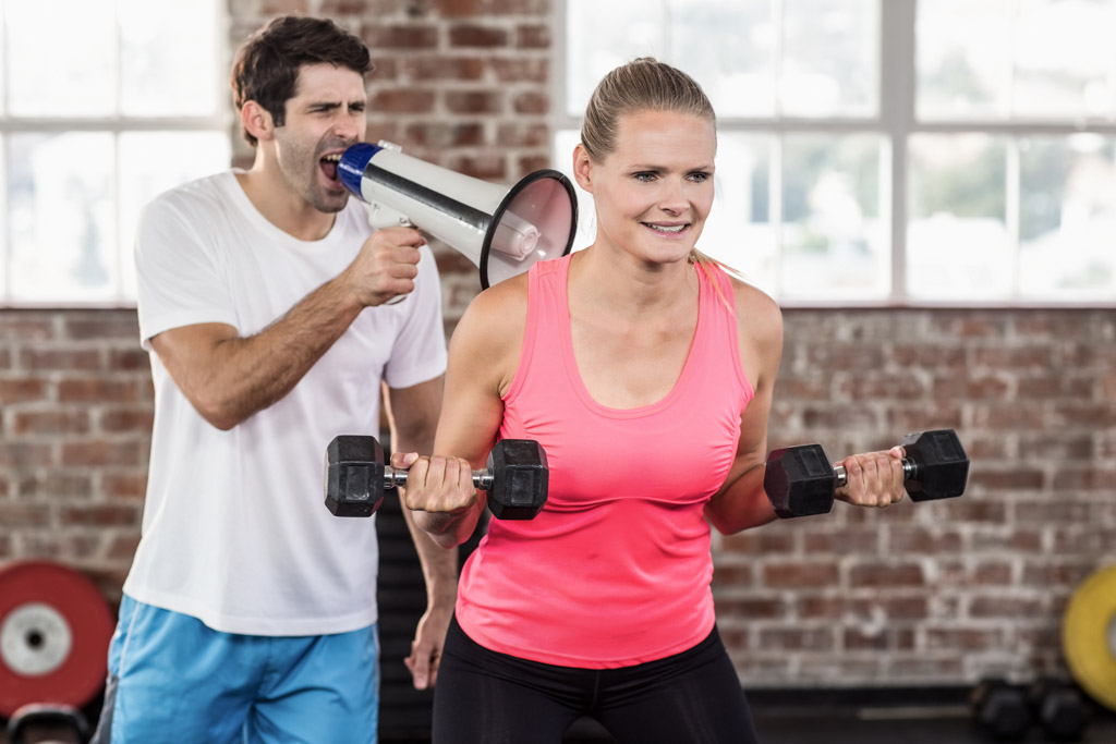 A trainer uses a bullhorn to yell at a client who is doing biceps curls in a gym.