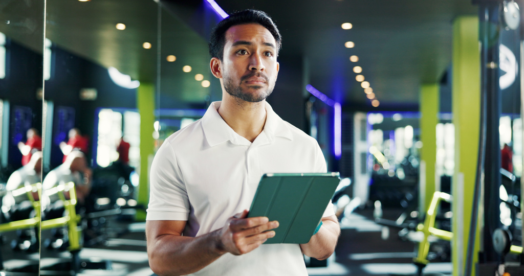 A stressed gym owner looks up from reviewing job applications on a tablet.